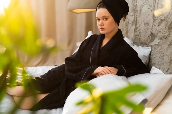 Siyah bornoz ve havlu turban takmış genç kadın, beyaz yastıklar üzerinde bitkiler önünde doğal ışıkta lifestyle çekimi.