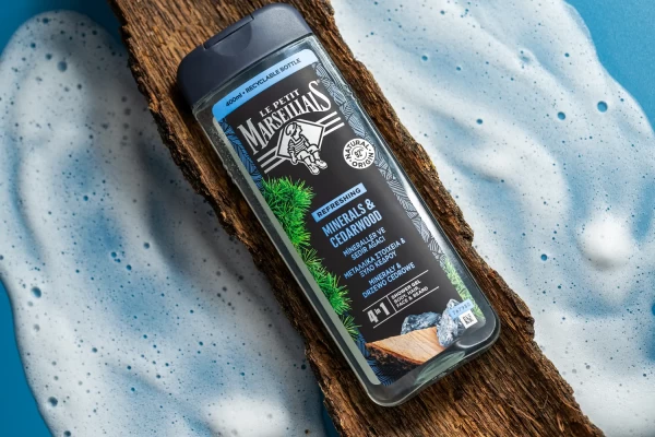 Şeffaf plastik şişedeki siyah-mavi cedarlı duş jeli, ağaç kabuğu üzerinde köpüklü fonda, detay odaklı ürün çekimi.