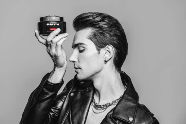 Siyah beyaz stüdyo portresinde deri ceketli erkek model alnında saç jeli kavanozu tutuyor, modern konsept kozmetik çekimi.