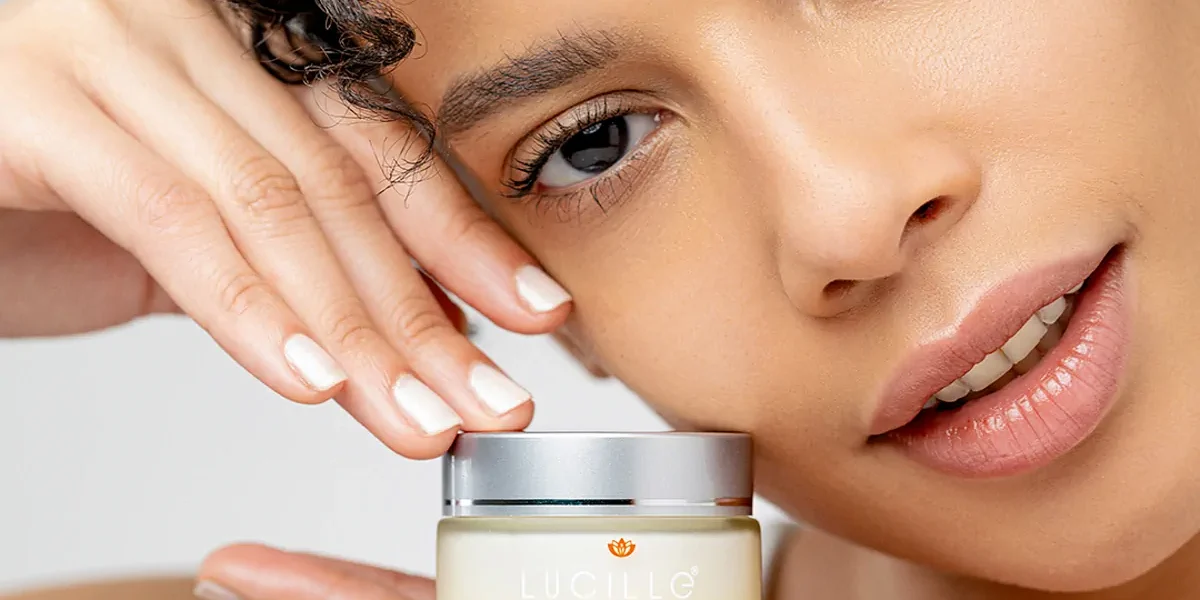 Yakın plan portrede model, beyaz fonda küçük Lucille markalı anti-aging krem kavanozunu tutuyor; doğal ışıkta kozmetik çekimi.
