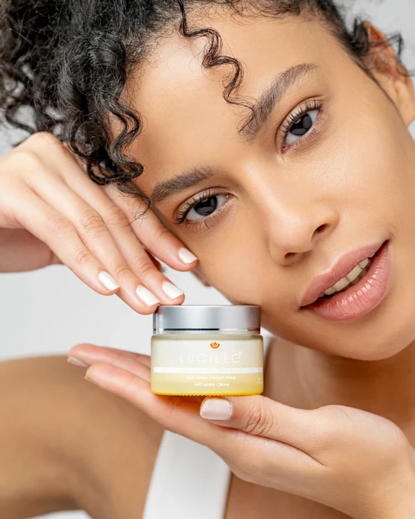 Yakın plan portrede model, beyaz fonda küçük Lucille markalı anti-aging krem kavanozunu tutuyor; doğal ışıkta kozmetik çekimi.