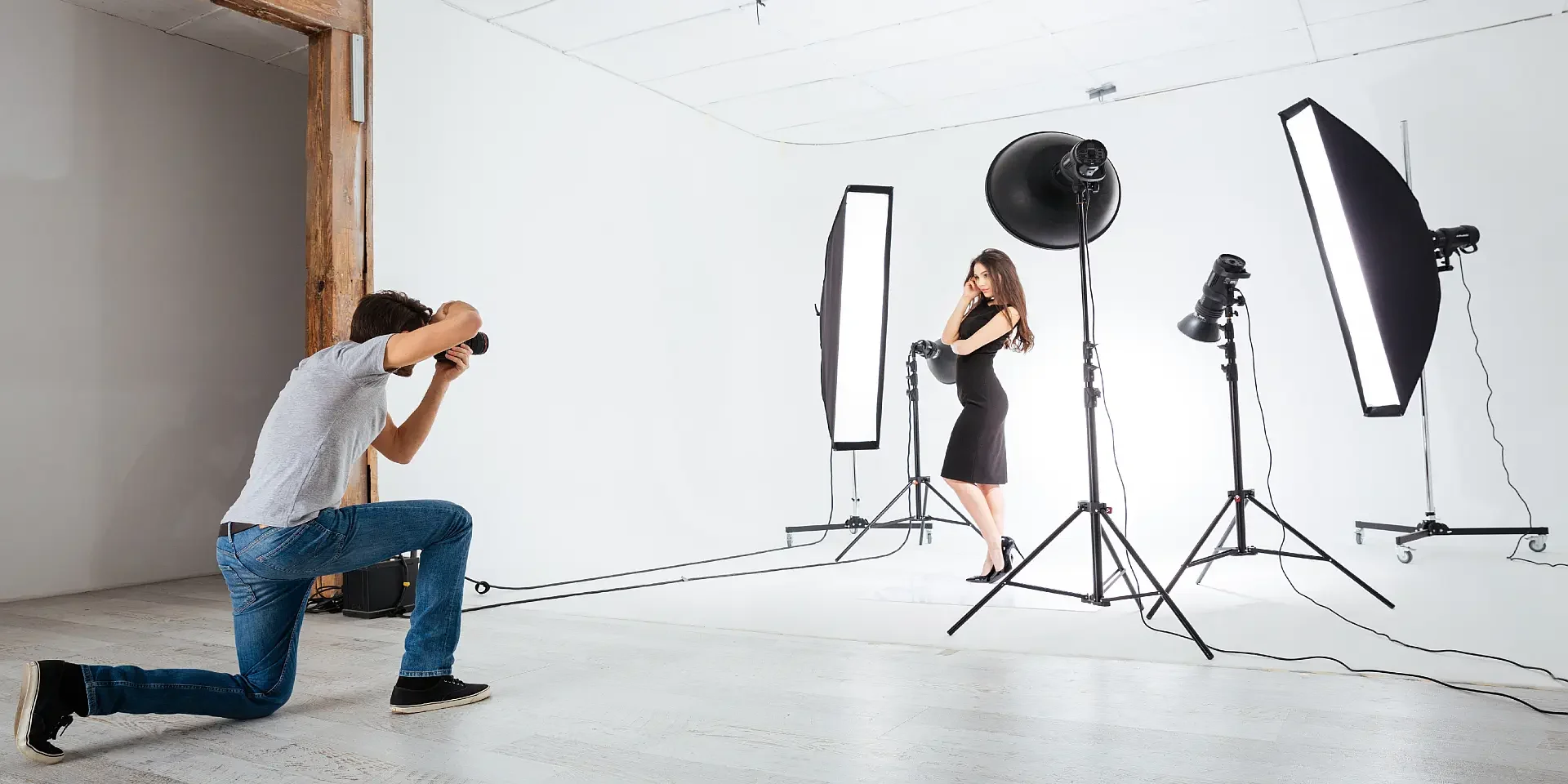Beyaz stüdyo fonunda softbox ışıklarla çevrili siyah elbiseli mankeni diz çökmüş fotoğrafçı çekiyor, modern mankenli çekim.