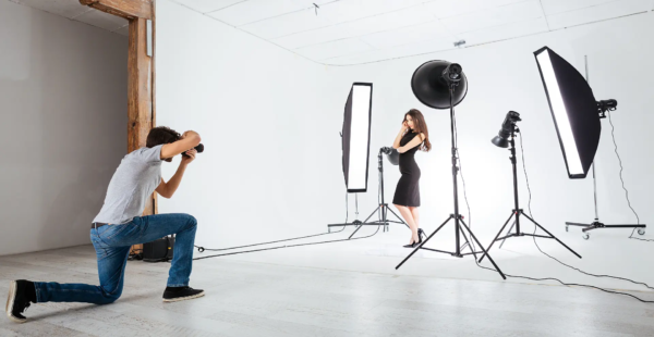 Beyaz stüdyo fonunda softbox ışıklarla çevrili siyah elbiseli mankeni diz çökmüş fotoğrafçı çekiyor, modern mankenli çekim.