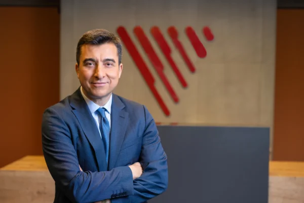 Koyu mavi takım elbiseli iş insanı, kolları çapraz, sıcak ofis fonunda kırmızı logo ile modern portre çekimi.