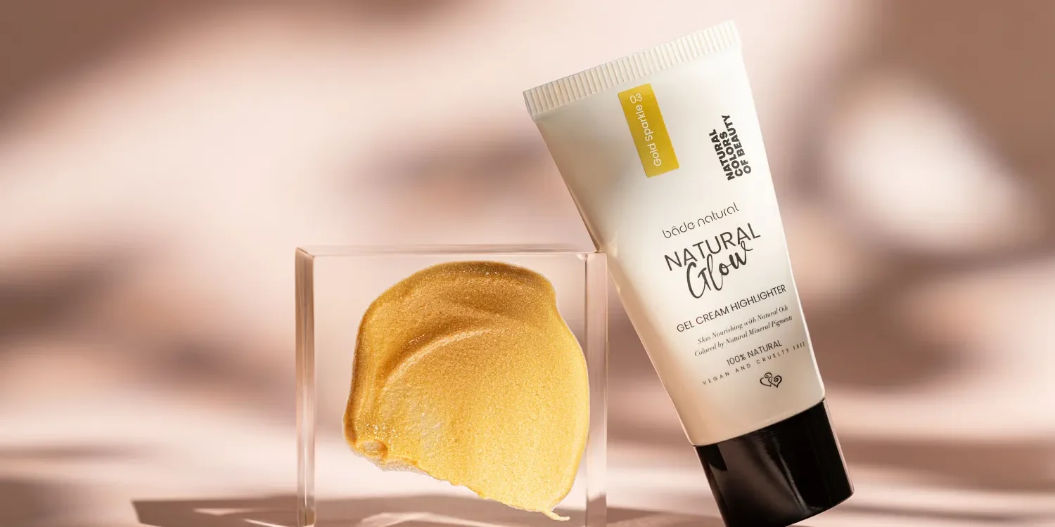 Sedefli altın highlighter kremi ve tüp ambalajı şeffaf blok üzerinde, yumuşak doğal ışıkta minimalist kozmetik çekimi.