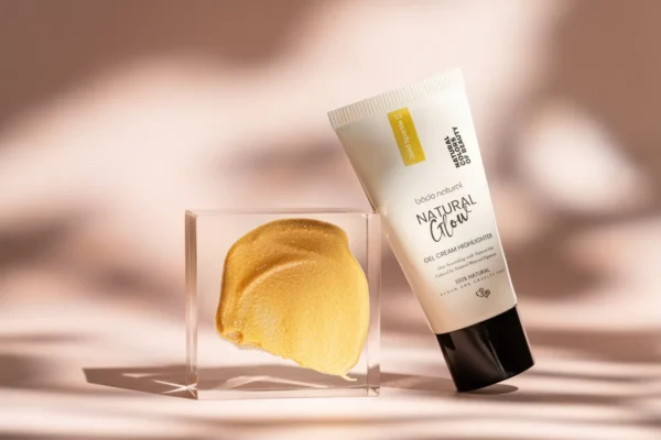 Sedefli altın highlighter kremi ve tüp ambalajı şeffaf blok üzerinde, yumuşak doğal ışıkta minimalist kozmetik çekimi.