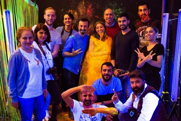Creative studio shot with woman wrapped in yellow fabric and smiling crew around her in studio covered with splashes of colorful paint.