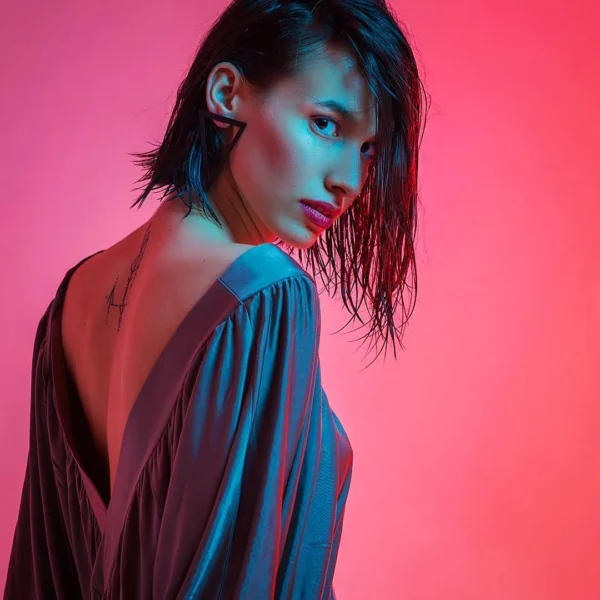 Modern editorial shot of woman with wet hair wearing backless purple dress, blue accent lights on pink gradient background.