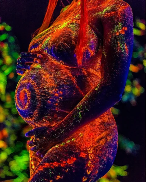 Detailed, conceptual studio portrait shot of a pregnant woman's abdomen and chest covered in neon UV paints