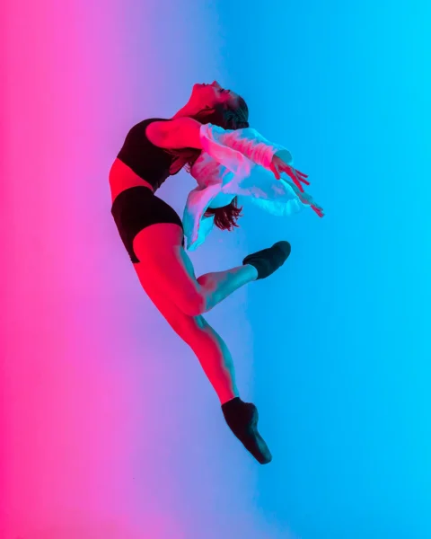 Modern studio shot of female dancer in black sportswear and light white shirt leaping in the air against neon pink-blue gradient background.