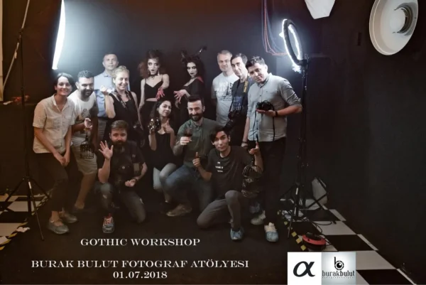 Two models with gothic make-up and their team pose together in a studio setting against a black background; concept portrait shoot.