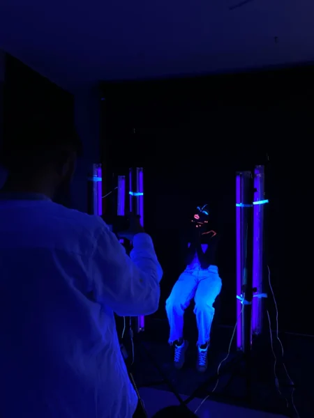 Photographer shooting a sitting model with neon face paint under purple-blue UV lights in a dark studio, concept studio shoot