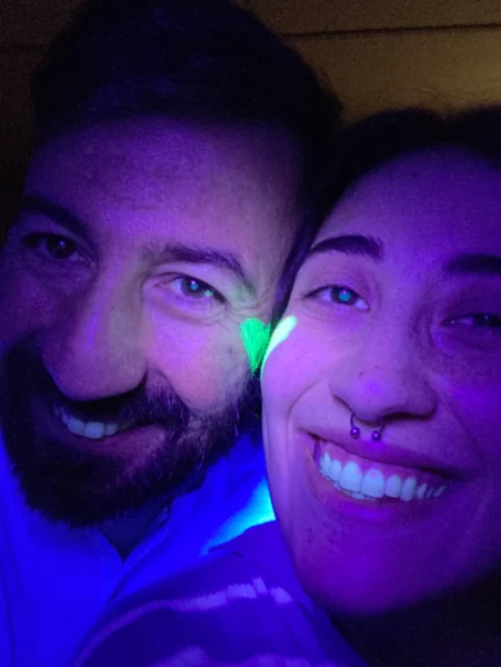 Concept portrait shot with smiling couple in close-up in purple-blue light, green heart paint on the nose and septum piercing.