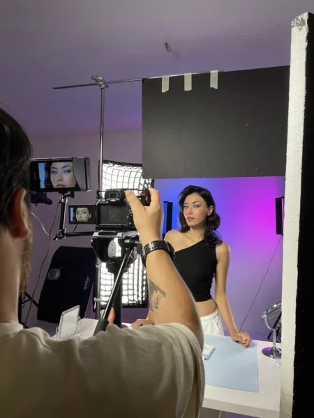 Photographer using light and camera to capture the model wearing a black one-shoulder top in front of a purple-pink background; modern studio shot.