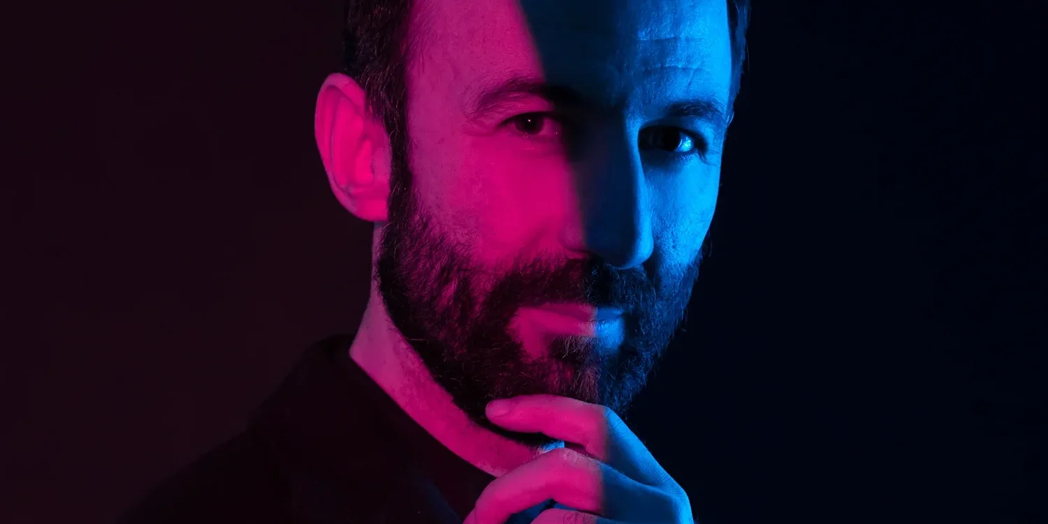 Karanlık fonda sakallı bir adam, yüzü pembe ve mavi ışıkla bölünmüş şekilde çenesini tutuyor; modern portre çekimi.