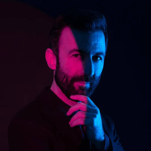 Karanlık fonda sakallı bir adam, yüzü pembe ve mavi ışıkla bölünmüş şekilde çenesini tutuyor; modern portre çekimi.