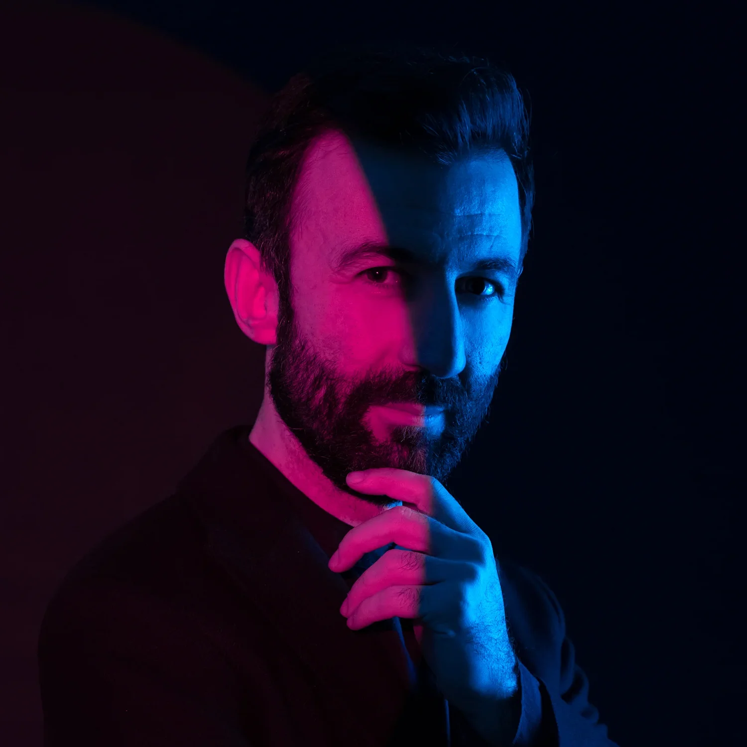 Karanlık fonda sakallı bir adam, yüzü pembe ve mavi ışıkla bölünmüş şekilde çenesini tutuyor; modern portre çekimi.