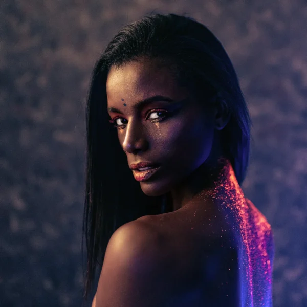 Studio portrait shot of dark-skinned woman in side profile, dramatic eye makeup, and pink sparkles on her neck, modern.