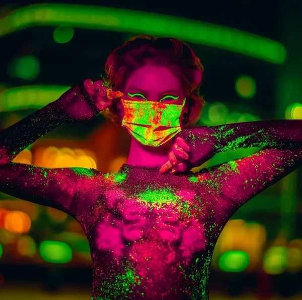 UV ışık altında pembe ve neon yeşil parlayan vücut boyasıyla maskeli kadın, şehir ışıkları önünde konsept editorial çekim.
