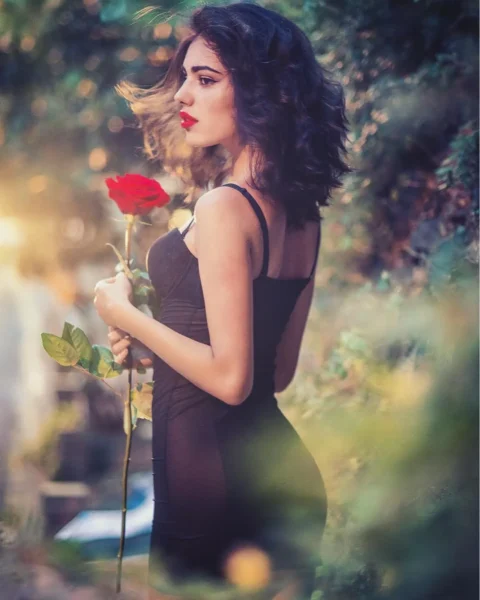 Yan profilden siyah elbise giymiş genç kadın kırmızı bir gülü tutuyor, flu doğal ışıklı arka planda romantik editorial çekim.