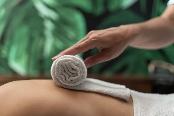Sırtın üzerinde beyaz rulo havlu ve uzanan elin bulunduğu spa sahnesi, doğal ışıkta lifestyle çekimi atmosferi.