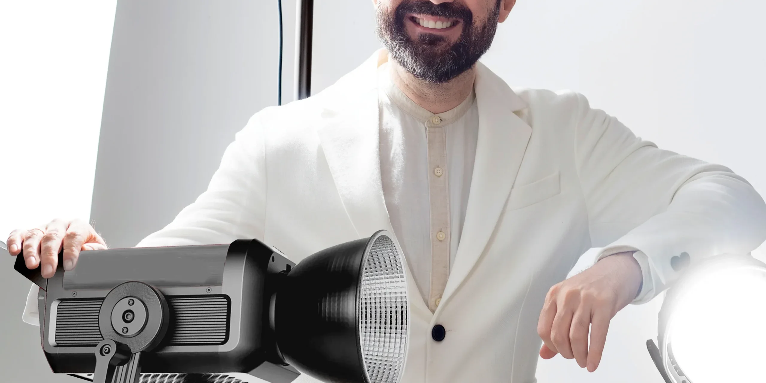 Beyaz ceketli sakallı adam, parlak stüdyo ışıkları önünde açık fonda gülümseyerek poz veriyor; modern stüdyo çekimi.