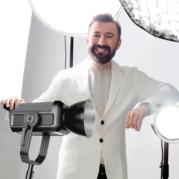 Beyaz ceketli sakallı adam, parlak stüdyo ışıkları önünde açık fonda gülümseyerek poz veriyor; modern stüdyo çekimi.