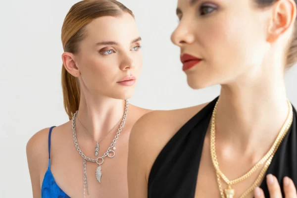 Beyaz fon önünde iki modelin gümüş ve altın kolyelerini vurgulayan minimalist takı çekimi, yakın plan ve temiz kompozisyon.